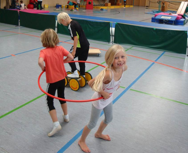 Kinder spielen mit einem Reifen und einem Pedalo in der Sporthalle.